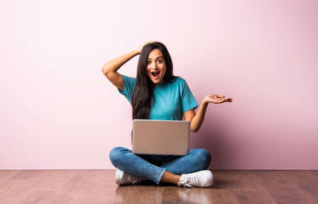 Indian asian young woman or girl sitting with laptop on her lap against pink wall on wooden floorの写真素材