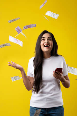 Happy Indian woman using smartphone and flying or raining or falling money, Rupee banknotes on...