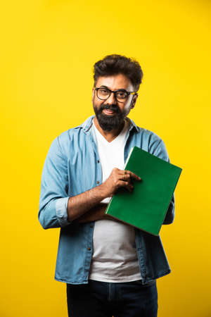 Young Indian happy man wearing eyeglasses reading book and smiling isolated over yellow background