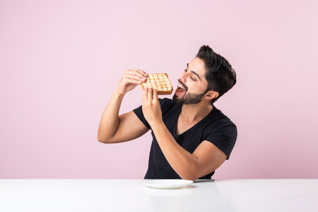 Indian Asian young bearded man eats bread sandwich while sitting in kitchen or dining table. Showing or presentingの写真素材