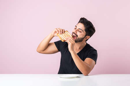 Indian Asian young bearded man eats bread sandwich while sitting in kitchen or dining table. Showing...