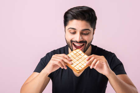 Indian Asian young bearded man eats bread sandwich while sitting in kitchen or dining table. Showing or presentingの写真素材