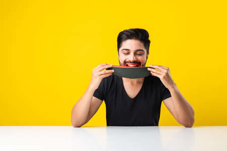 Portrait of a handsome Indian young man smiling and eating fresh watermelon or tarbooj while sitting at table or isolated over wooden floorの写真素材