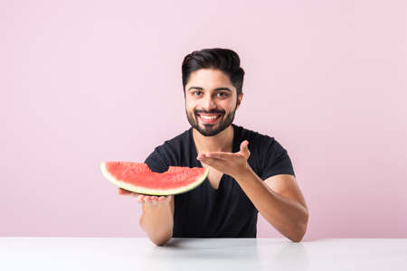 Portrait of a handsome Indian young man smiling and eating fresh watermelon or tarbooj while sitting at table or isolated over wooden floorの写真素材
