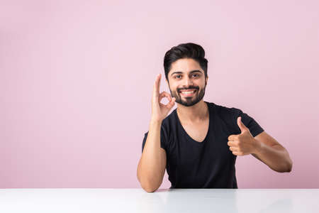 Successful Indian young man celebrating victory with thumbs up sign, sitting at desk or table...