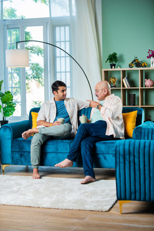 Happy Indian father and son having tea or coffee while sitting on sofa in living roomの写真素材