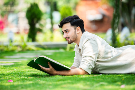 Young attentive Indian male student lies on green grass and reads book at summer green parkの写真素材