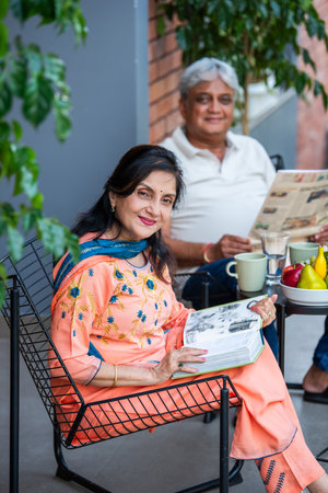 Elderly Indian asian couple reading newspaper and book while listning to music and having breakfast outdoors in galleryの写真素材