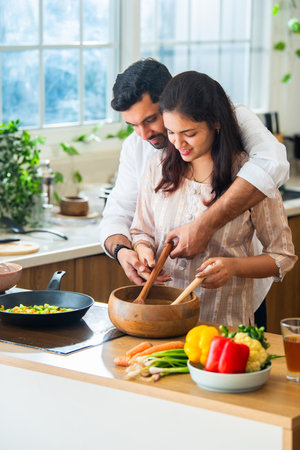 Happy loving Indian asian young couple cooking food in kitchen together, hugging, standing at tableの写真素材