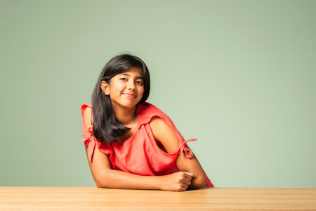 Portrait of Indian asian small girl sitting at table against green wallの写真素材