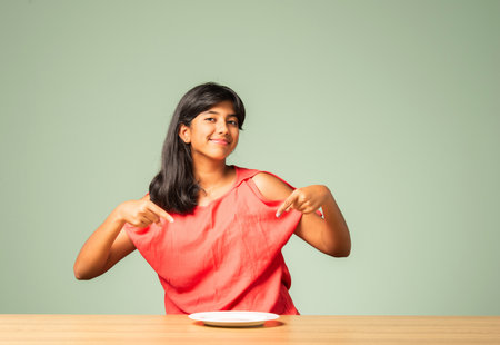 Indian asian small girl presenting empty plate while sitting at tableの写真素材