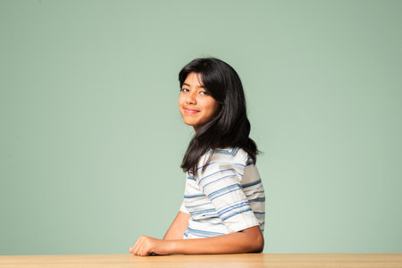 Portrait of Indian asian small girl sitting at table against green wallの写真素材