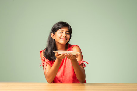 Indian asian small girl presenting empty plate while sitting at tableの写真素材