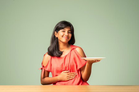 Indian asian small girl presenting empty plate while sitting at tableの写真素材