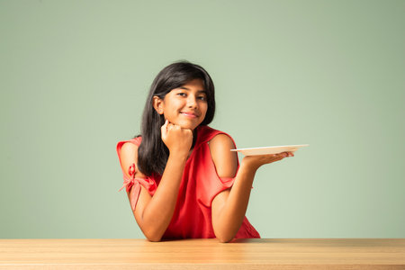 Indian asian small girl presenting empty plate while sitting at tableの写真素材