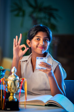 Indian asian small girl child drinking milk in cup or glass while studying at homeの写真素材