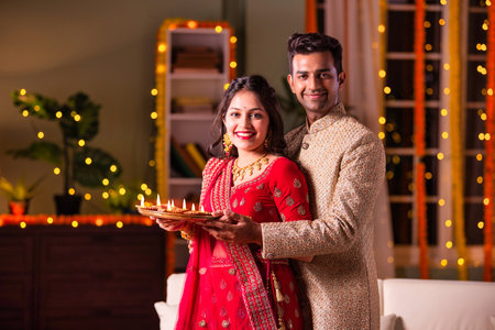 Indian married young couple celebrating diwali festival holding puja thali and sweets while sitting on sofaの写真素材