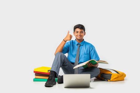 Indian Schoolboy in uniform with books and laptop, pointing at blank space, promoting educationの写真素材