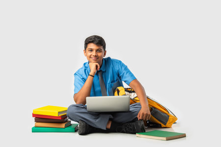 Indian Schoolboy in uniform with books and laptop, pointing at blank space, promoting educationの写真素材