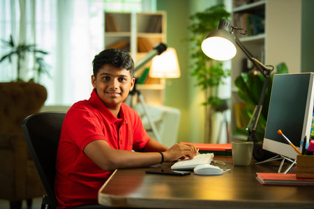 Indian Asian teenage boy studying using computer, headphones, smartphone for online class, homeworkの写真素材