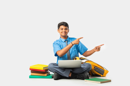 Indian Schoolboy in uniform with books and laptop, pointing at blank space, promoting educationの写真素材