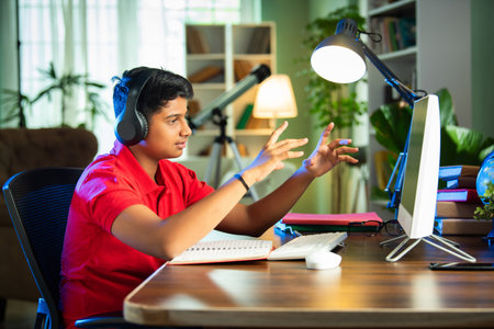 Indian Asian teenage boy studying using computer, headphones, smartphone for online class, homeworkの写真素材