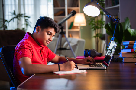 Indian Asian teenage boy studying using computer, headphones, smartphone for online class, homeworkの写真素材