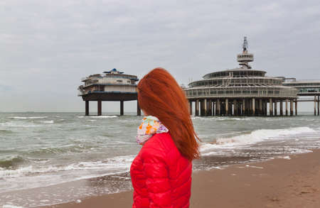Read haired woman is looking to waving sea and pier. Woman is wearing in red jacket.の写真素材