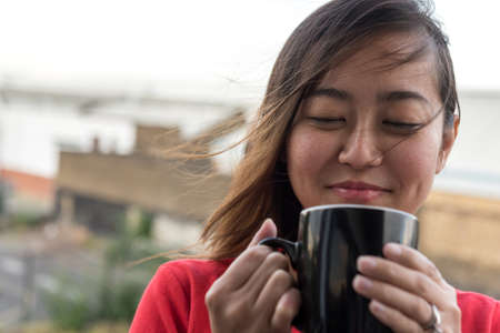 Hip Young Asian Woman Drinking Cup of Coffee on Balcony with Urban Background Sceneの写真素材