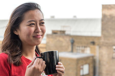 Hip Young Asian Woman Drinking Cup of Coffee on Balcony with Urban Background Scene