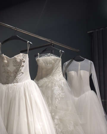 Selection of handmade white wedding gowns hanging on a rail in a darkened room partially illuminated by a lightの写真素材