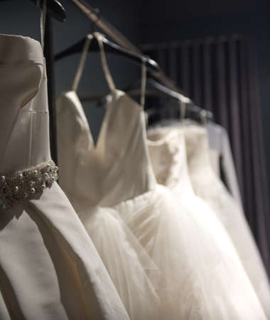 Selection of handmade white wedding gowns hanging on a rail in a darkened room partially illuminated by a lightの写真素材