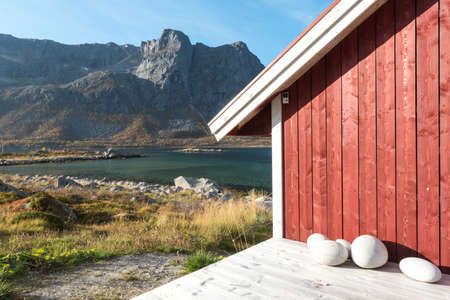 Close up cropped view of a wooden Norwegian beach hut with stones on the shoreline overlooking the Tromso Fjord, Arctic Circle, Norway and mountain peak on a sunny blue sky dayの写真素材