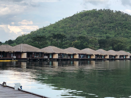 Floating accommodations with thatched roofs line a calm body of water at a resort in Kanchanaburi, Thailand.の写真素材