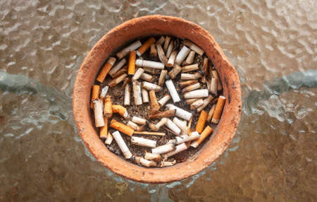 Ashtray full of burnt cigarette butts, made of clay and over a glass table.の写真素材