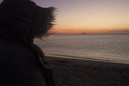 Woman in the beach watching sea in winter, wearing a coat, with the hood on, during sunset and there is a ship.の写真素材
