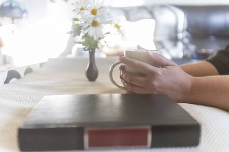 Woman taking a cup of coffee and a book on the table, a jar with flowers, daisies. Sunny day indoorの写真素材
