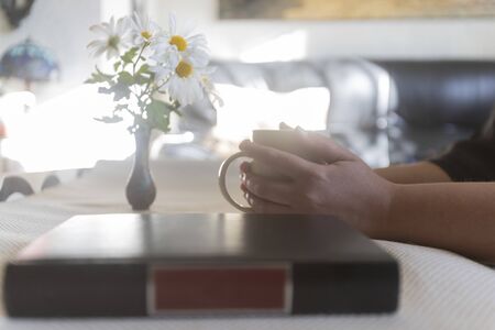 Woman taking a cup of coffee and a book on the table, a jar with flowers, daisies. Sunny day indoorの写真素材