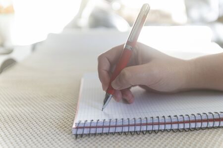 Young woman hand writing with a red pen on a notebook during a sunny day in a room at homeの写真素材