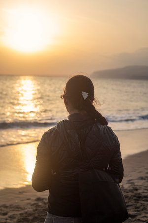 Photography of young and white woman looking at the sea during the sunset, with a braid and coat, during the winter.の写真素材