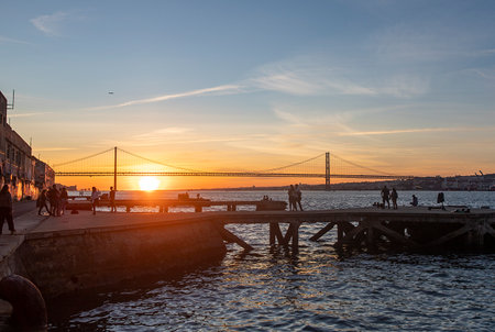 People watching the sunset in Cacilhas and Lisbon's April 25 Bridge, dock on the Tagus River and plane in the skyの写真素材
