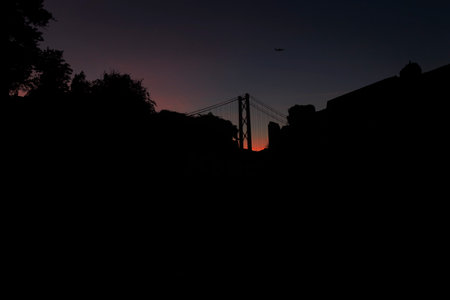 Silhouette photography of the 25 de Abril Bridge in Lisbon and plane in the sky, during the night, landscape with treesの写真素材