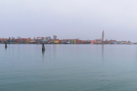 A stunning view of the coastline of Burano Island, seen from Mazzorbo, showcasing the iconic colorful houses and the leaning Campanile of San Martino Church. The serene setting highlights the unique charm of Venetian island life.の写真素材