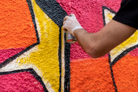 A close-up image showing a man's hand painting graffiti on a wall. The spray can release vibrant...