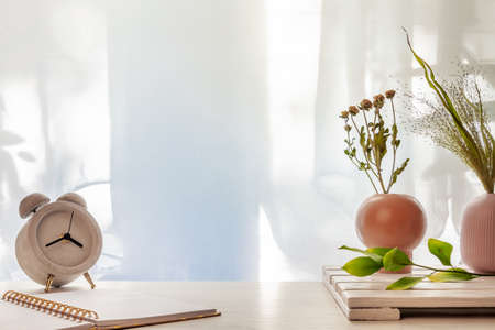 Photo of a desk with a concrete clock, an open notebook and vases with dried grasses on a background of sun-lit curtainsの写真素材