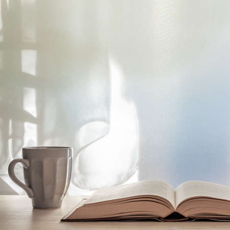 Photo of a desk with an open book and a gray cup with tea on a background of sun-lit curtains. Copy spaceの写真素材