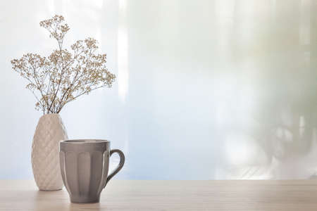 A photo of a desk with a white vase with dried white flowers and a gray cup with coffee on a background of sun-lit curtains. Copy spaceの写真素材