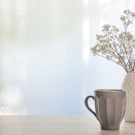 A photo of a wooden desk with a white vase with dried white flowers and a gray cup with tea on a background of sun-lit curtains. Copy spaceの写真素材