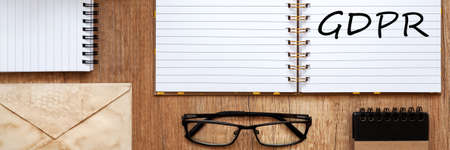 Panoramic view on wooden desk with open notebook with GDPR text, old envelope and black glasses.