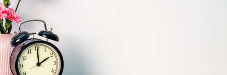 A panoramic photo of white empty copy space wall with black alarm clock and pink carnations in a...